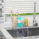 A clear plastic SuoXing sink caddy with multiple compartments holding sponges, a dish brush, and soap bottles.